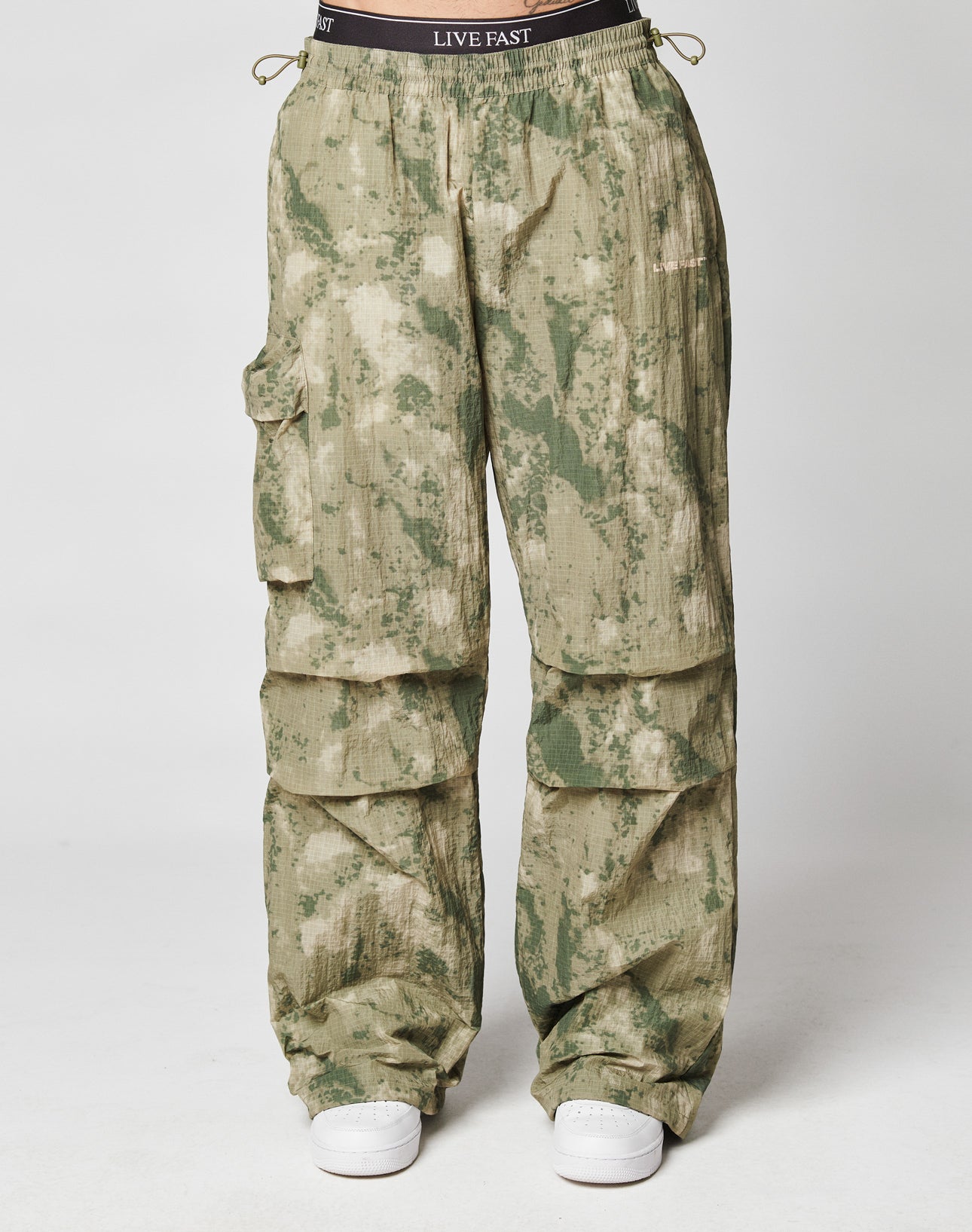 The LIVE FAST DIE YOUNG Live Fast Nylon Pants feature a loose oversize fit, elastic cord ankles, green/beige camo print, and a waistband with LIVE FAST print. Shown with white sneakers on a plain white background.