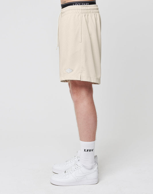 A person, shown from the waist down, models LIVE FAST DIE YOUNG Basic Mesh Shorts in beige with a logo patch, paired with white LFDY crew socks and white Nike sneakers against a plain white background.