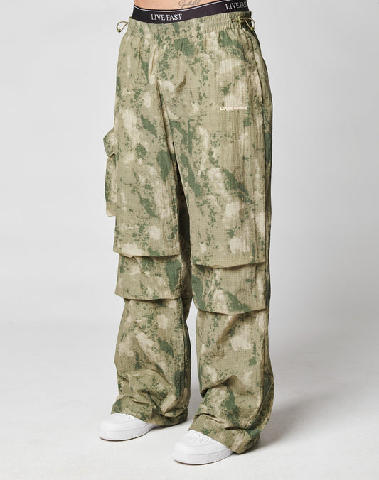 Wearing LIVE FAST DIE YOUNG’s Live Fast Nylon Pants in green camo with an oversize fit, multiple cargo pockets, elastic drawcords, and a “LIVE FAST” waistband print, styled with white sneakers against a plain white background.