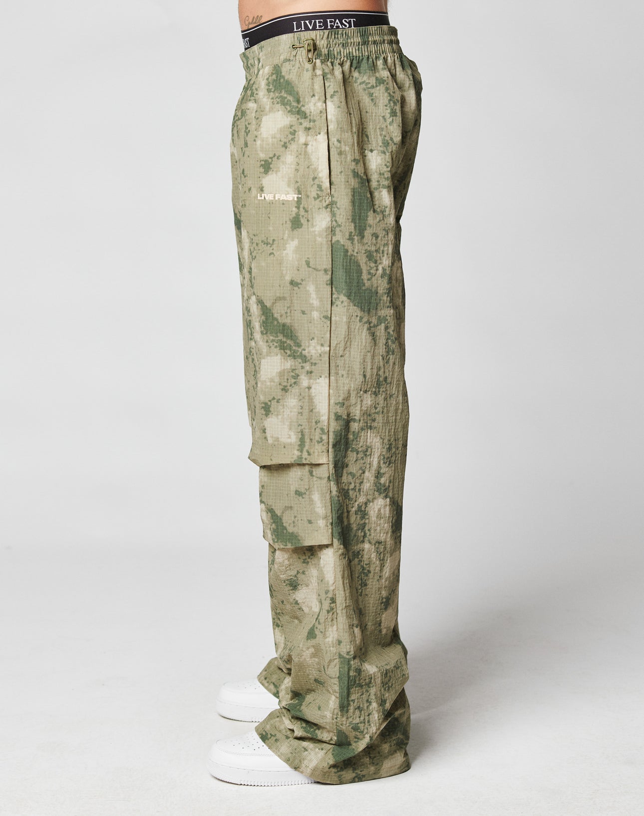 A person wearing LIVE FAST DIE YOUNGs Live Fast Nylon Pants in green camo with cargo pockets and LIVE FAST on the waistband and pocket, stands sideways in white sneakers against a plain white backdrop.
