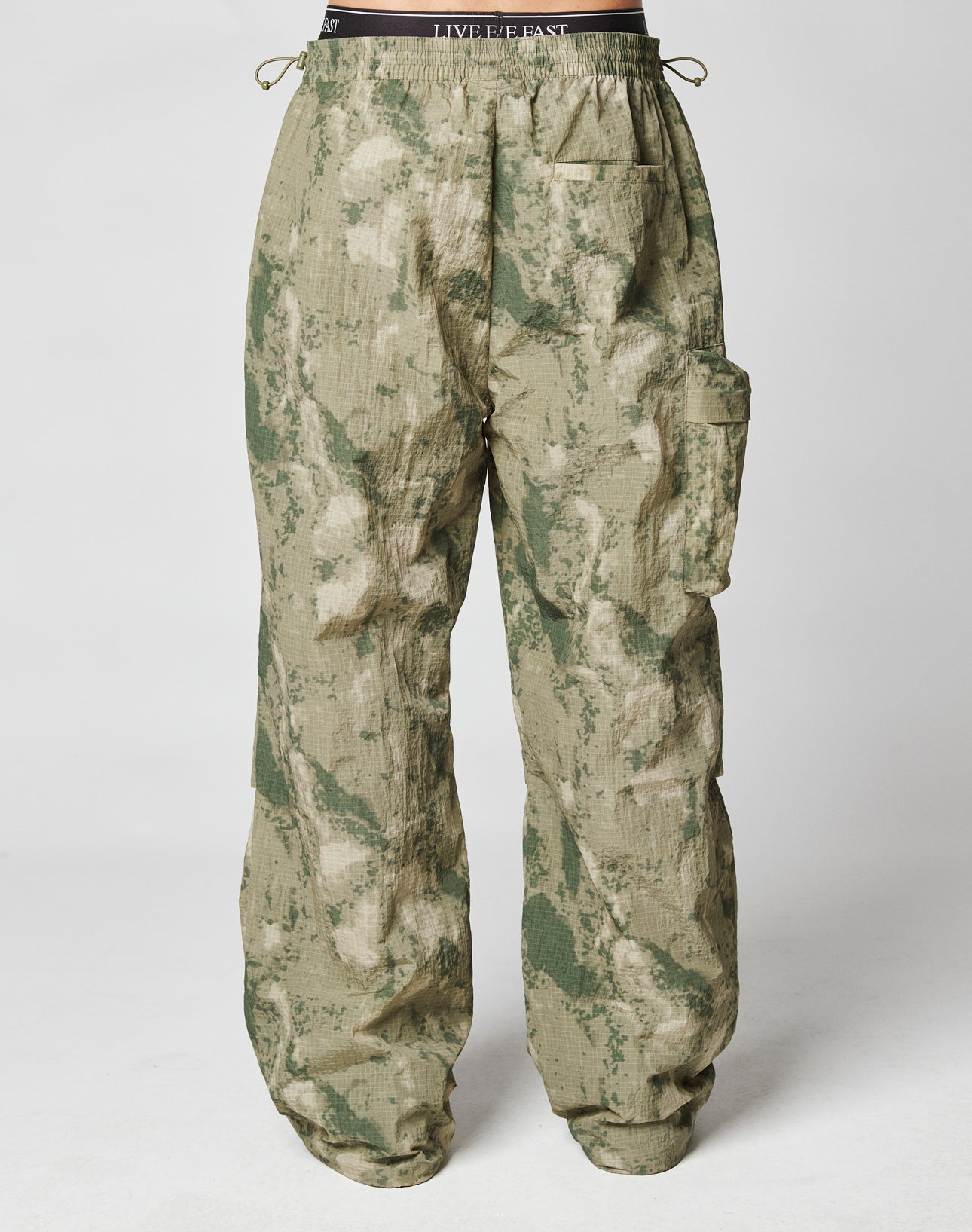 The Live Fast Nylon Pants by LIVE FAST DIE YOUNG are loose-fit green and tan camo cargo pants with Cargotasche details, an elastic waistband, and side pockets, shown on a model against a white background.