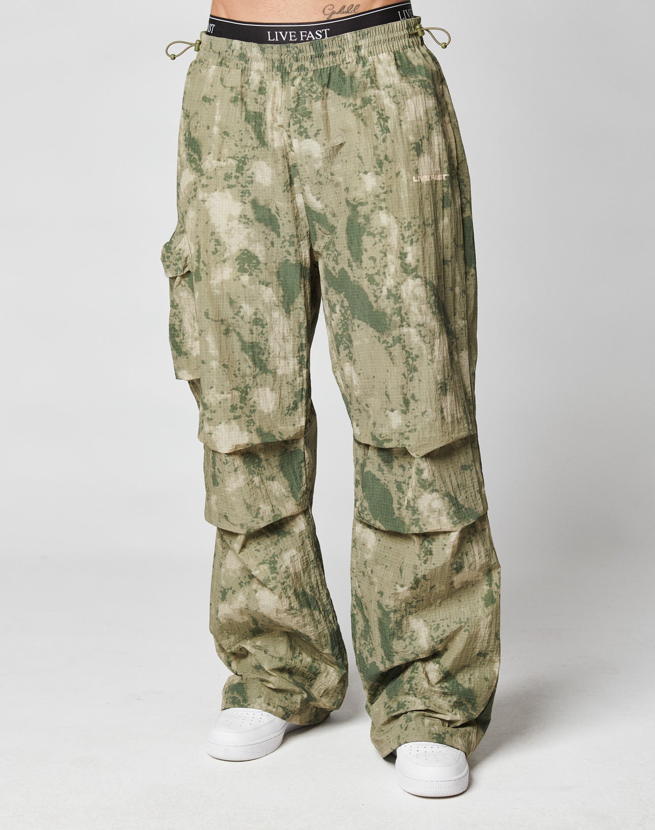 A person stands against a white background in LIVE FAST DIE YOUNG Live Fast Nylon Pants—oversized green and beige camouflage cargos with an elastic waistband, LIVE FAST printed on it, multiple cargo pockets, and paired with white sneakers.