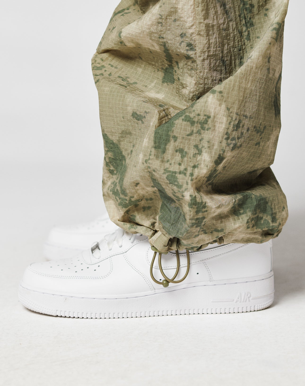 A close-up side view of a person wearing LIVE FAST DIE YOUNG Live Fast Nylon Pants in green camouflage with drawstring waist and cargo pockets, paired with white Nike Air Force 1 sneakers against a plain white background.