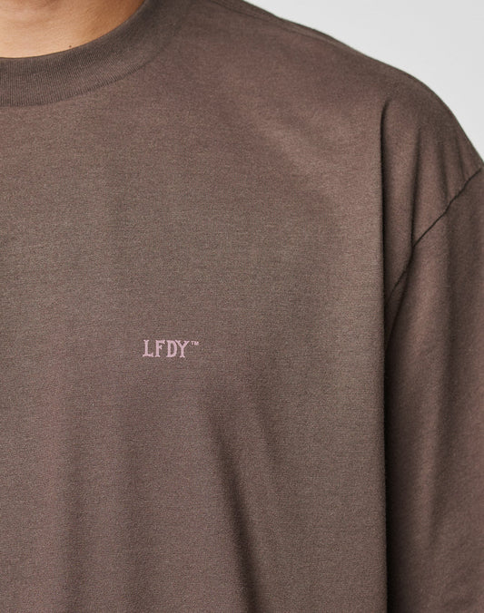 A close-up of a person in the LFDY Flash Tee by LIVE FAST DIE YOUNG—an oversize brown crewneck T-shirt, crafted from soft Cotton-Slub Stoff, with “LFDY™” embroidered in small light pink lettering on the chest.