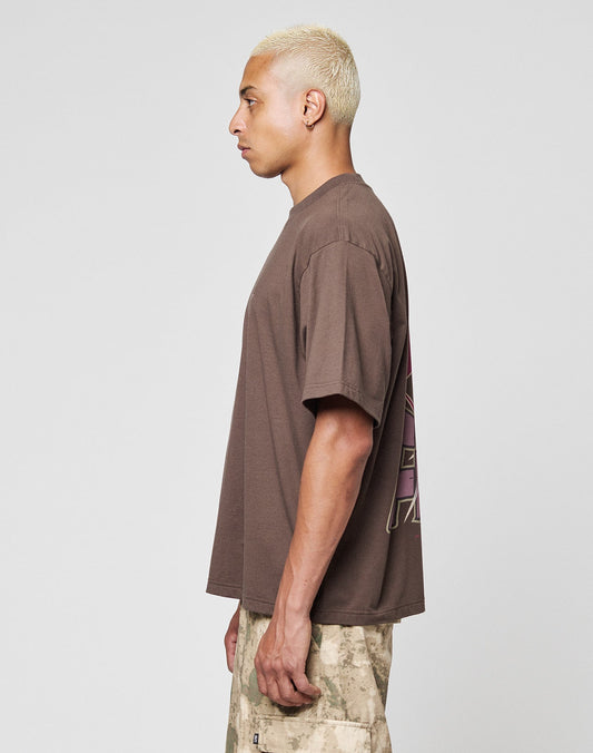 A person with short bleached hair stands in profile, wearing a loose brown LFDY Flash Tee from LIVE FAST DIE YOUNG and beige camouflage shorts against a plain light gray background.