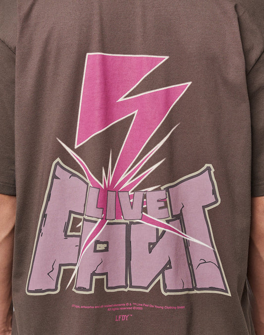 A person wears the LFDY Flash Tee by LIVE FAST DIE YOUNG—an oversized brown Cotton-Slub shirt with a large pink lightning bolt and bold LIVE FAST prints front and back, featuring a dynamic cracked effect around the text.