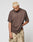 A person with short bleached blond hair wears a loose LFDY Flash Tee by LIVE FAST DIE YOUNG and tan camouflage cargo pants, standing against a plain light background and looking down while gripping their shirt.