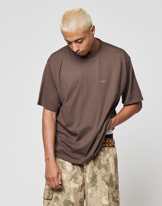 A person with short bleached blond hair wears a loose LFDY Flash Tee by LIVE FAST DIE YOUNG and tan camouflage cargo pants, standing against a plain light background and looking down while gripping their shirt.