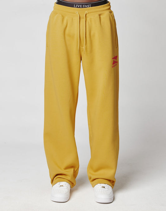 A person wears the LIVE FAST DIE YOUNG Live Fast Sweatsuit Jogger in mustard yellow with an oversize fit, elastic waistband, red logo on the left thigh, white sneakers, and a black LIVE FAST waistband against a plain white background.