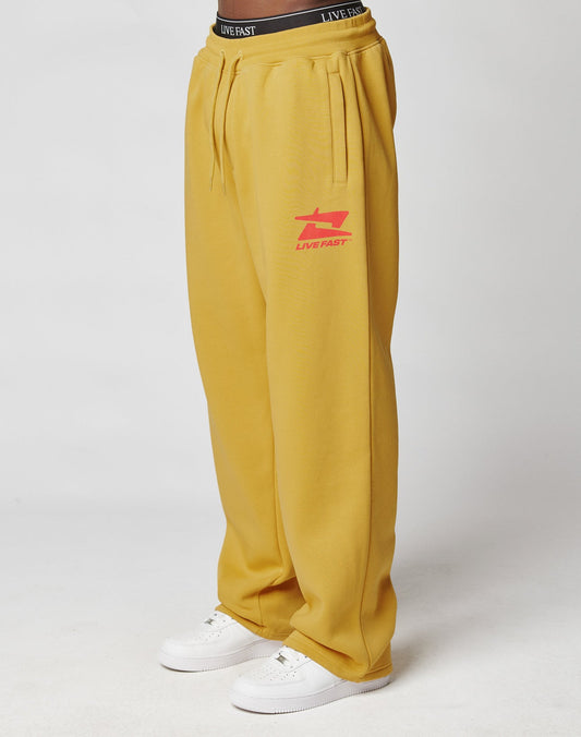 A person wears the LIVE FAST DIE YOUNG Live Fast Sweatsuit Jogger in mustard yellow with a red LIVE FAST logo on the left thigh, Elastik-Bund, oversize fit, white sneakers, and a visible black waistband. Background is plain white. | model