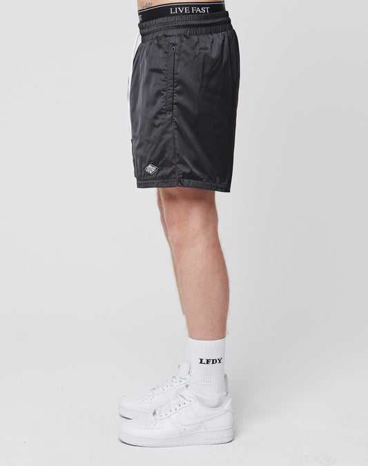 A person stands sideways wearing LIVE FAST DIE YOUNG Basic Satin Shorts featuring a logo patch and “LIVE FAST” on the elastic waistband, paired with white “LFDY” socks and white sneakers, against a plain white background.