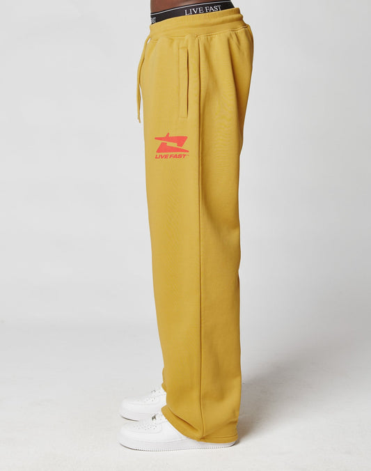A person models the LIVE FAST DIE YOUNG Live Fast Sweatsuit Jogger in mustard yellow with an elastic waistband and red LIVE FAST logo, styled in an oversize fit with white sneakers. Only the lower body is shown against a white background.