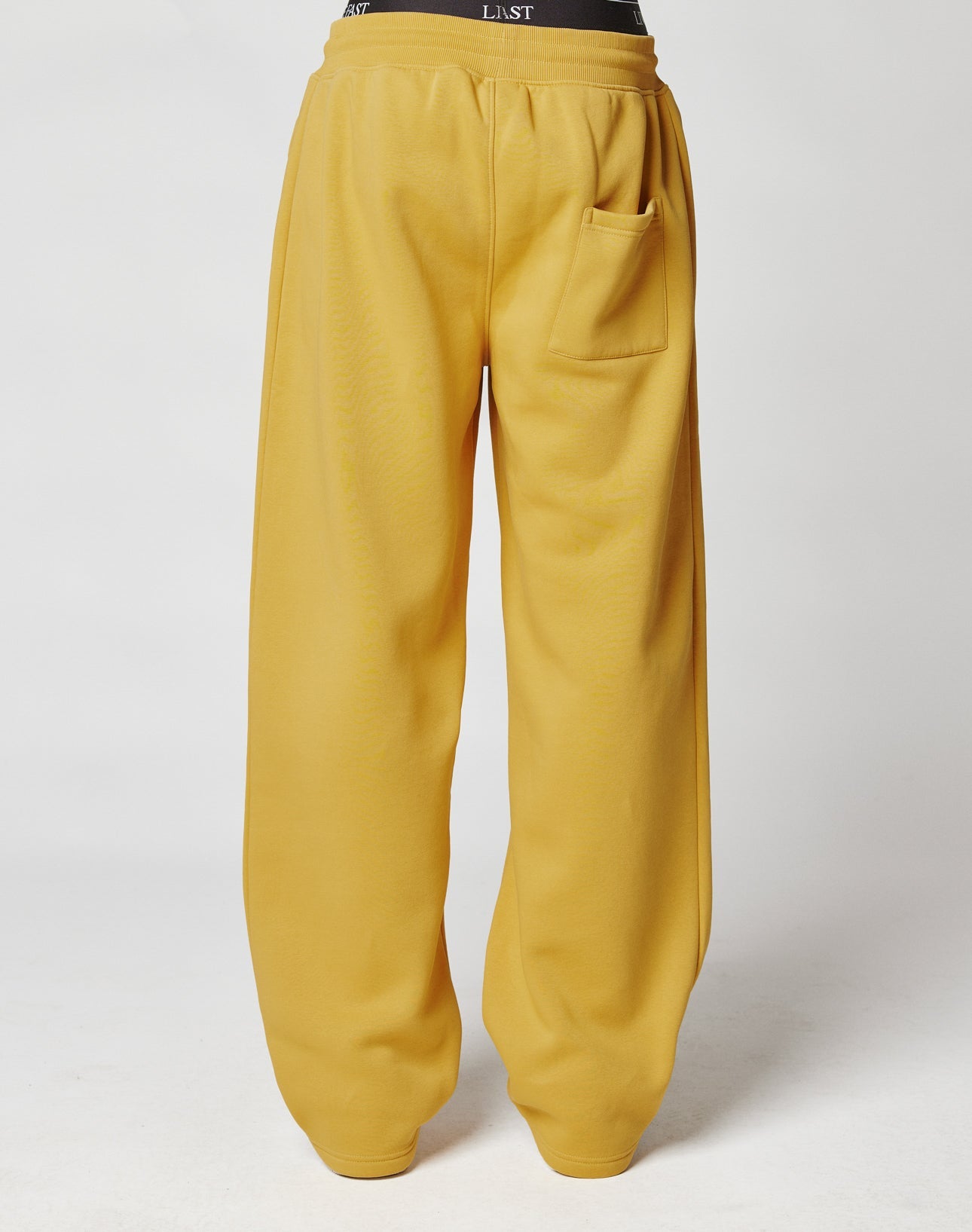 The Live Fast Sweatsuit Jogger by LIVE FAST DIE YOUNG is a loose-fitting mustard yellow sweatpant with an oversize fit, single back pocket, elastic waistband, and black label. Made from soft cotton. Shown from the back on a plain white background.