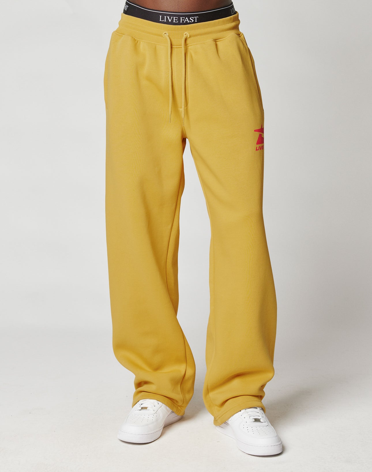 A person wears the LIVE FAST DIE YOUNG Live Fast Sweatsuit Jogger in mustard yellow Baumwolle with an oversize fit, white sneakers, and a black waistband featuring “LIVE FAST” in white, standing against a plain light background.