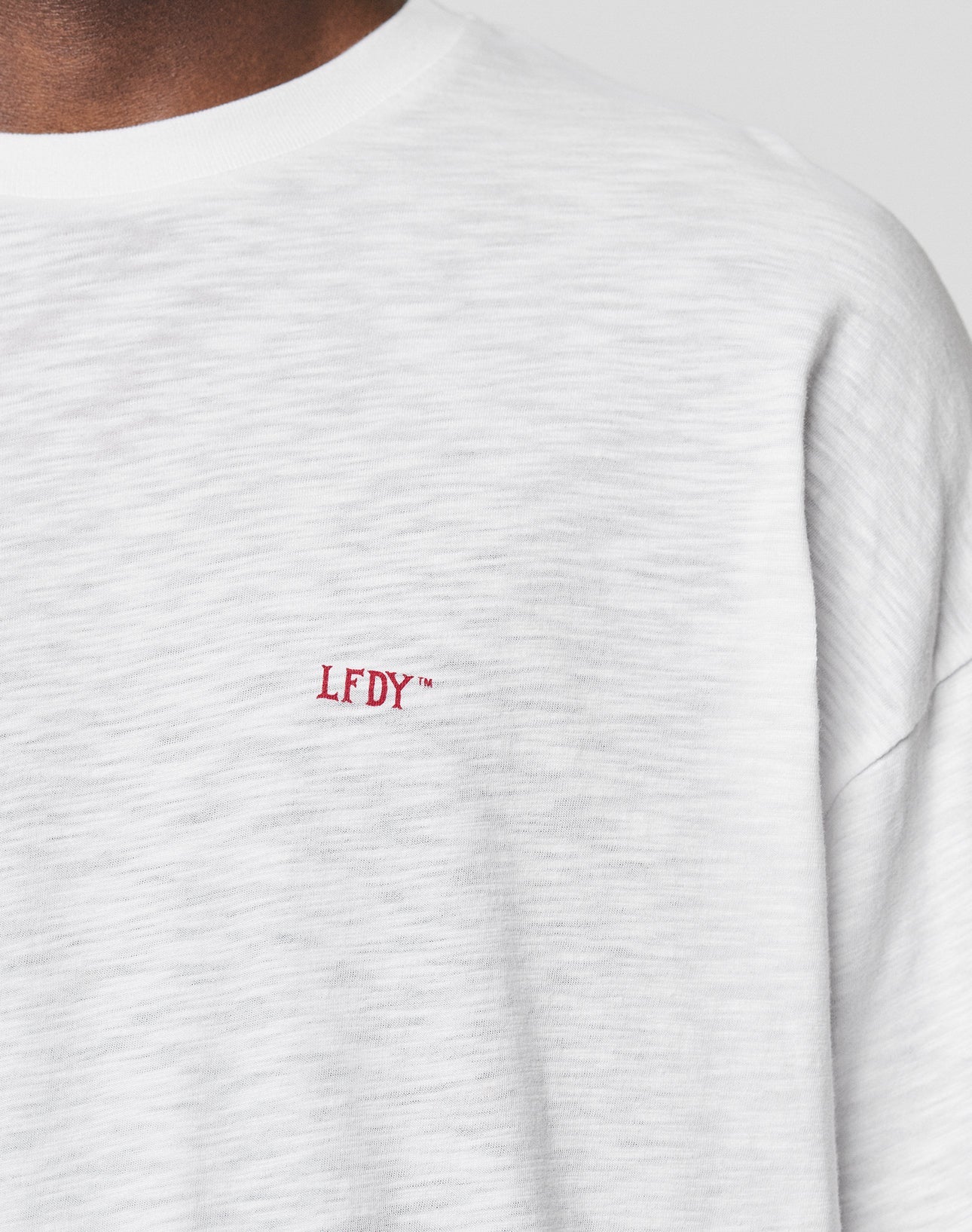 Close-up of a person wearing the LIVE FAST DIE YOUNG LFDY We Walked Longsleeve in white cotton-slub, featuring small red embroidered LFDY™ text on the left chest and showing part of their neck and shoulder.