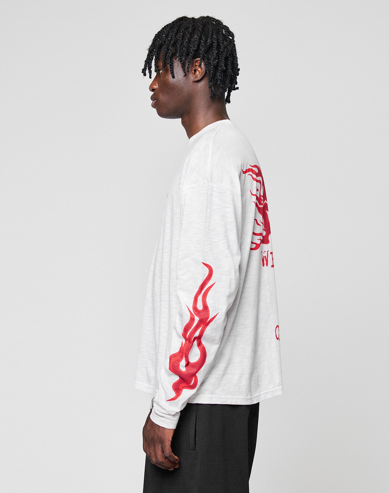 A person with short, twisted hair stands sideways in an oversized LIVE FAST DIE YOUNG LFDY We Walked Longsleeve featuring red flame graphics on the sleeves and back, paired with black pants, set against a plain light gray background.