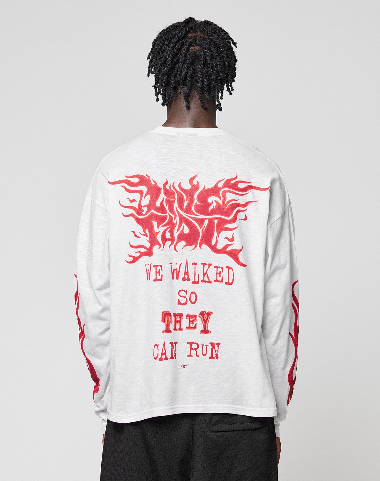 A person with braided hair, seen from behind, wears the LFDY We Walked Longsleeve by LIVE FAST DIE YOUNG—an oversized white 100% Baumwolle shirt with bold red “WE WALKED SO THEY CAN RUN.” text and flame designs on the back and sleeves.