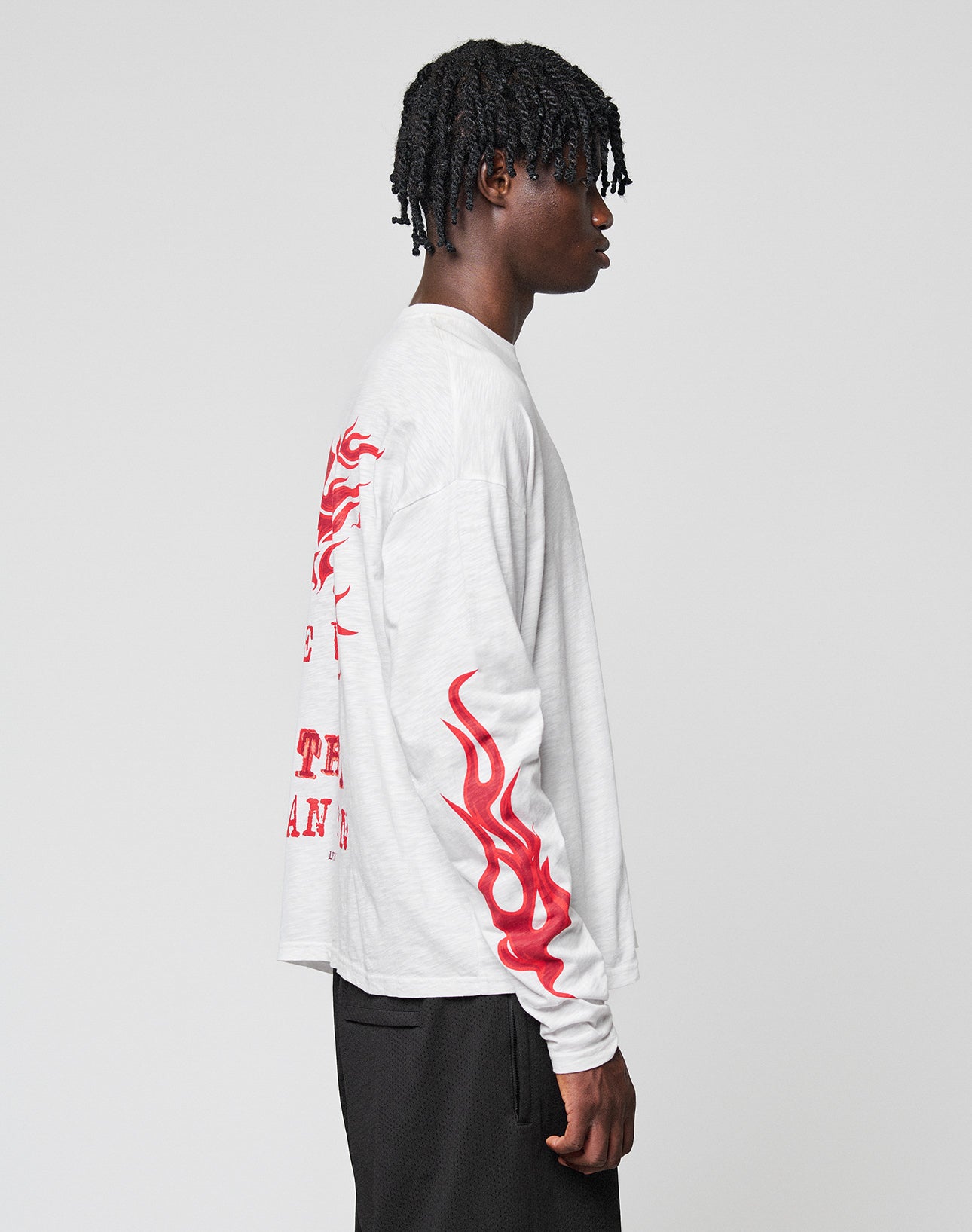 A person with short, twisted hair models the LIVE FAST DIE YOUNG LFDY We Walked Longsleeve—an oversized white shirt with red flame sleeve graphics and red back print—standing sideways against a plain light background.
