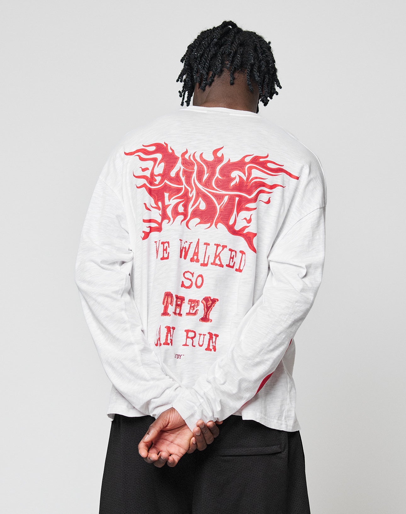 A person with short, twisted hair stands facing away, wearing the LIVE FAST DIE YOUNG LFDY We Walked Longsleeve—a 100% Baumwolle white shirt with bold red WE WALKED SO THEY CAN RUN text and a flame-like design above.