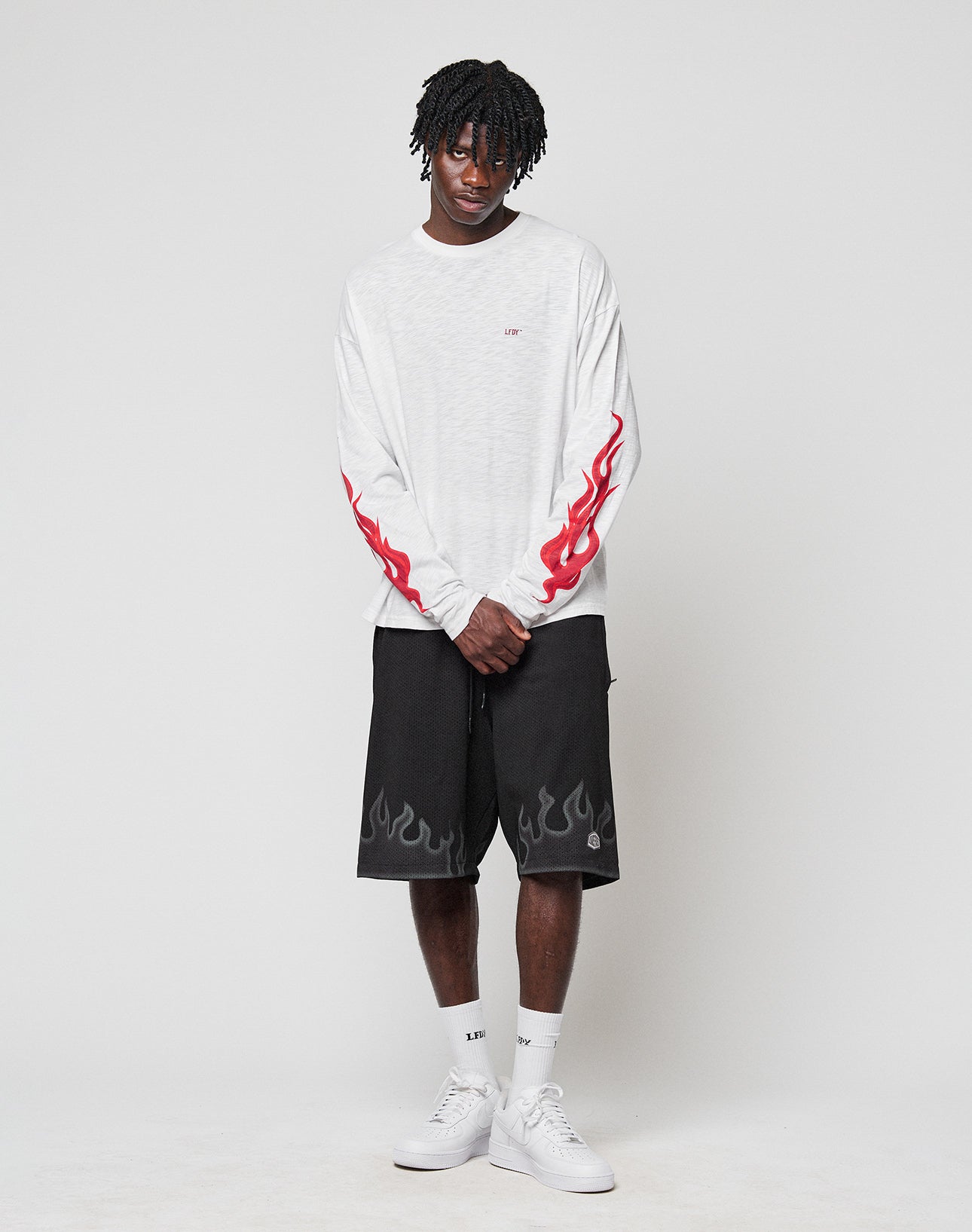 A young man stands against a plain background, wearing the LIVE FAST DIE YOUNG LFDY We Walked Longsleeve—an oversized white shirt with red flame sleeve designs—paired with black shorts, white socks, and white sneakers.