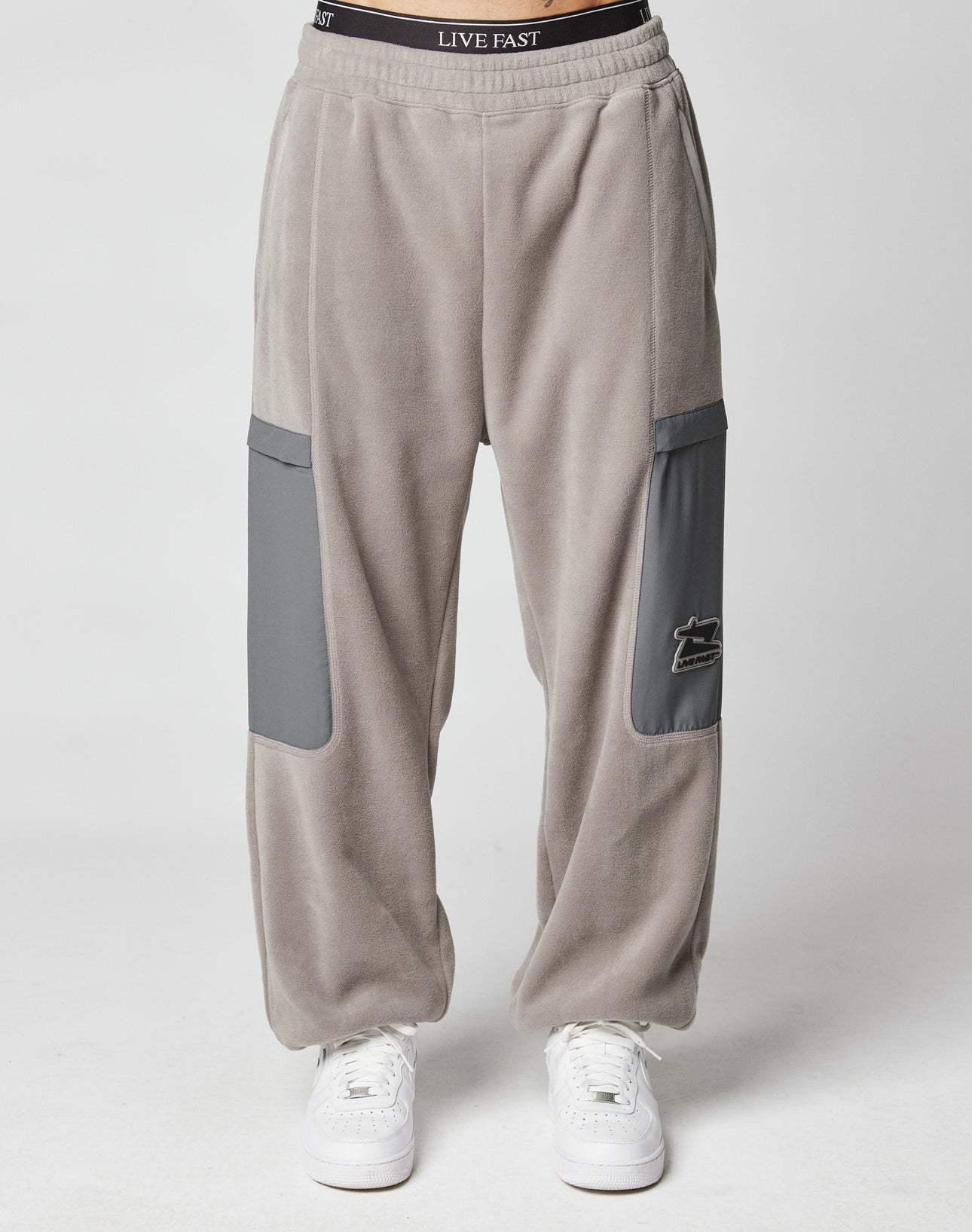 A person wears LIVE FAST DIE YOUNG’s Live Fast Fleece Pants in light gray with dark gray nylon thigh pockets, white sneakers, and a black waistband featuring LIVE FAST, standing before a plain white background.