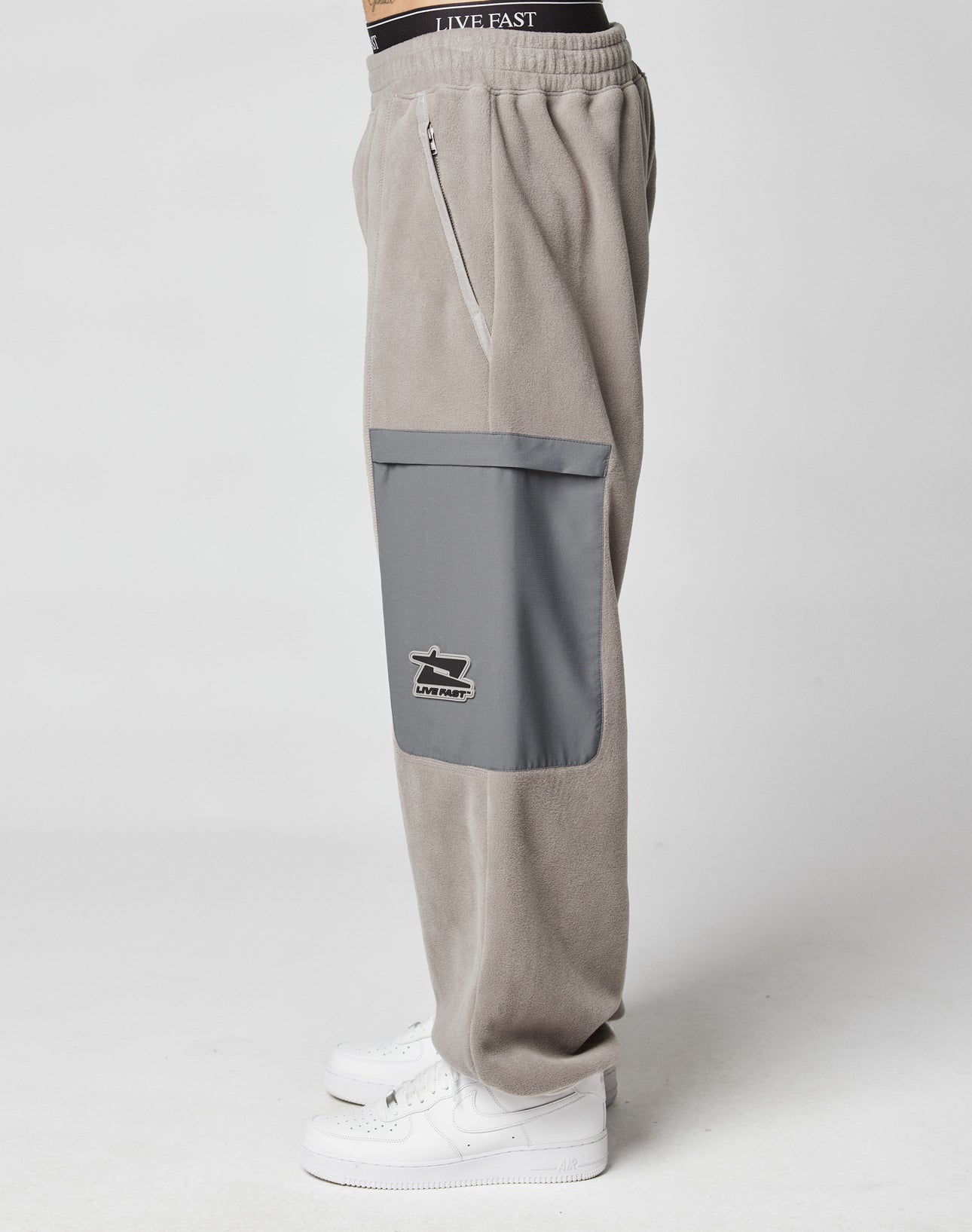 A person wearing LIVE FAST DIE YOUNG Live Fast Fleece Pants in light gray Wide Fit, featuring a darker gray nylon Taschen pocket and white logo patch, paired with white sneakers, shown from the side against a plain white background.