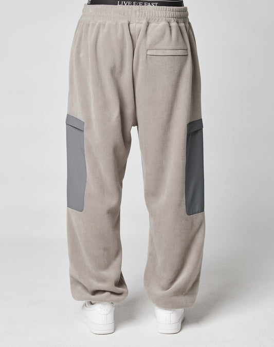A person is seen from behind wearing LIVE FAST DIE YOUNG Live Fast Fleece Pants in wide fit, light gray with dark gray Nylon Taschen patch pockets, an elastic waistband, and white sneakers, set against a plain light background.