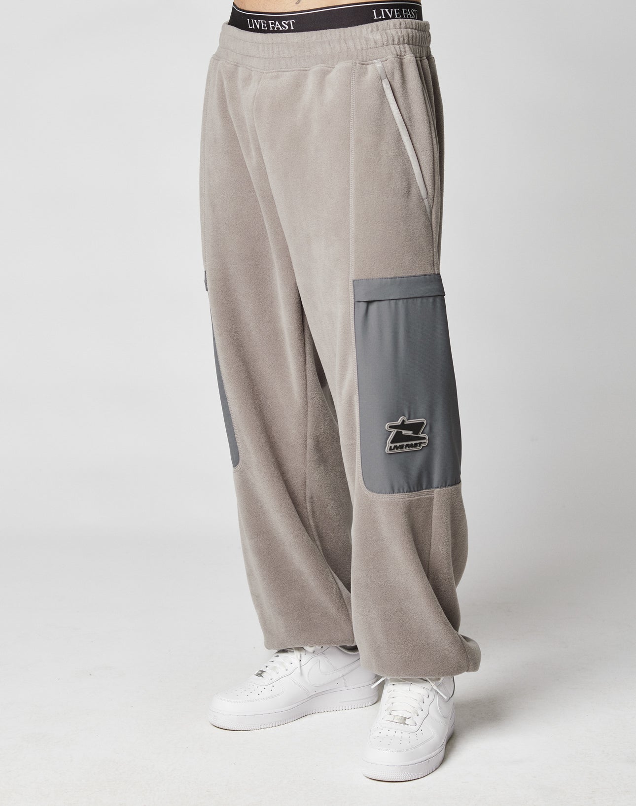 Person wearing LIVE FAST DIE YOUNG Live Fast Fleece Pants in wide fit gray with darker gray nylon pockets, a Live Fast logo, white sneakers, and an elastic waistband with LIVE FAST on a plain white background.