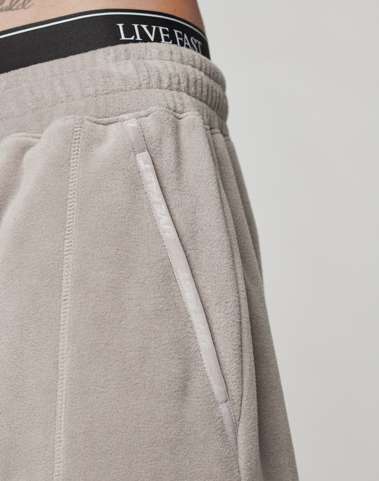 Close-up of a person wearing LIVE FAST DIE YOUNG Live Fast Fleece Pants in light gray, featuring an Elastik-Bund waistband with LIVE FAST in white, zippered pocket, Nylon Taschen, and a visible tattoo above the waistband.