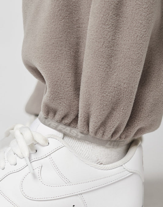 Close-up of a persons lower leg wearing LIVE FAST DIE YOUNG Live Fast Fleece Pants in beige with an elastic waistband and nylon pockets, white socks, and wide fit white lace-up sneakers.