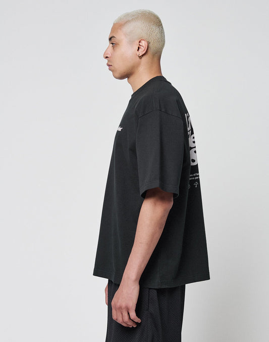A person with short, bleached blonde hair stands in profile against a plain background, wearing the LIVE FAST DIE YOUNG Basic 280 Tee in black and matching shorts. The shirt showcases both front and back print details.