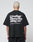 A person with short bleached hair wears a black LIVE FAST DIE YOUNG Basic 280 Tee aus Baumwolle, showcasing bold white “Live Fast Heavy Tee 280g/m²” text with Front- und Backprint icons. The background is plain and light-colored.