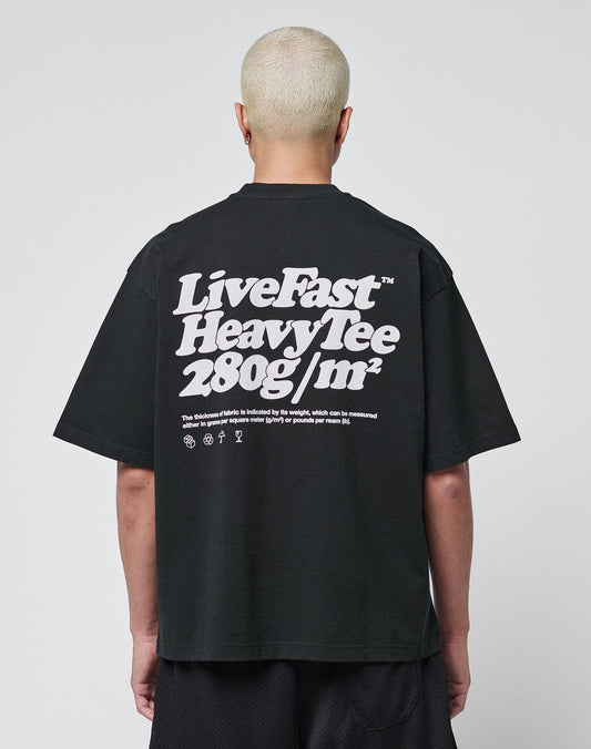 A person with short bleached hair wears a black LIVE FAST DIE YOUNG Basic 280 Tee aus Baumwolle, showcasing bold white “Live Fast Heavy Tee 280g/m²” text with Front- und Backprint icons. The background is plain and light-colored.