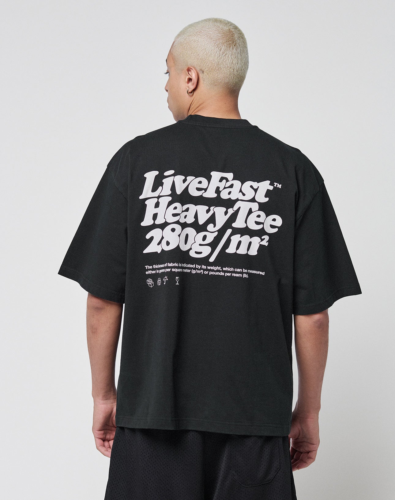 A person with short blond hair is seen from behind wearing the LIVE FAST DIE YOUNG Basic 280 Tee, an oversize black cotton T-shirt with bold white front and back prints and smaller text beneath.