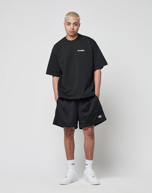 A person with short blond hair wears a LIVE FAST DIE YOUNG Basic 280 Tee in black with front and back print, black shorts, white socks, and white sneakers, standing against a plain light gray background facing the camera.