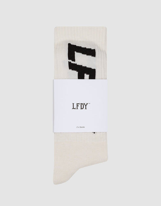 A 2-pack of off-white LIVE FAST DIE YOUNG Basic Typo Socks featuring a black geometric design, folded with a branded LFDY band labeled "2 x Socks" on a plain light gray background. Includes care instructions.