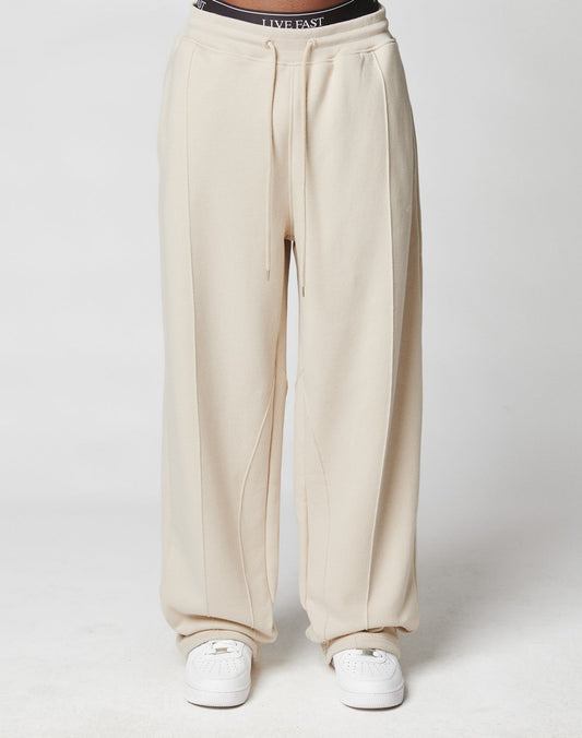 A person wears the LFDY Pleated Jogger by LIVE FAST DIE YOUNG in cream (wide fit, visible seams, elastic waistband, cotton), styled with white sneakers and black LIVE FAST underwear waistband peeking out. Set against a plain white background.