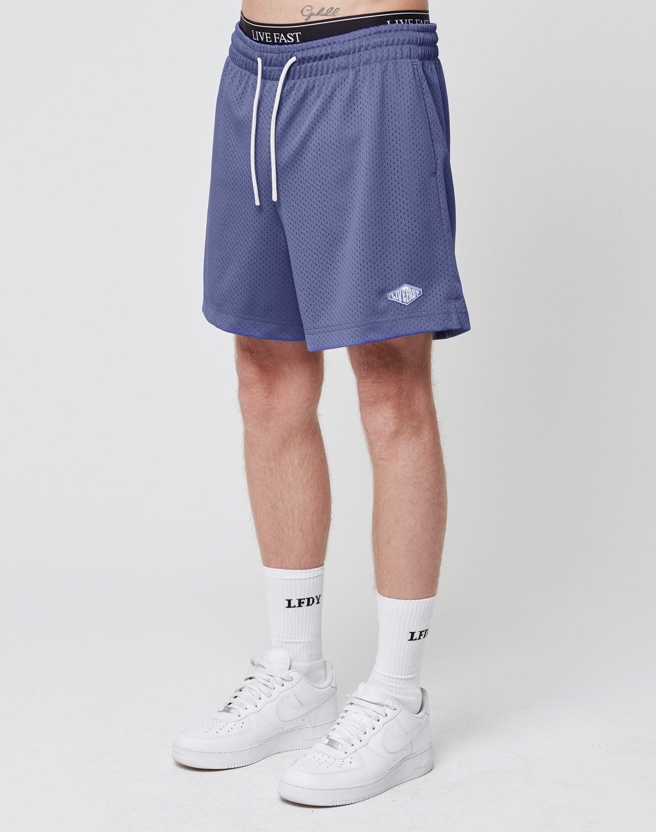 A person wearing LIVE FAST DIE YOUNG Basic Mesh Shorts in blue with an elastic waistband and regular fit, white LFDY socks, and white Nike sneakers stands against a plain white background. Only the lower body is shown. | model