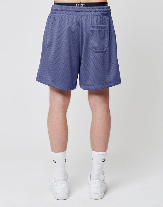 A person faces away, wearing LIVE FAST DIE YOUNG Basic Mesh Shorts in lavender with an elastic waistband and logo patch, plus a small back pocket. They pair them with white crew socks and sneakers against a plain white background.