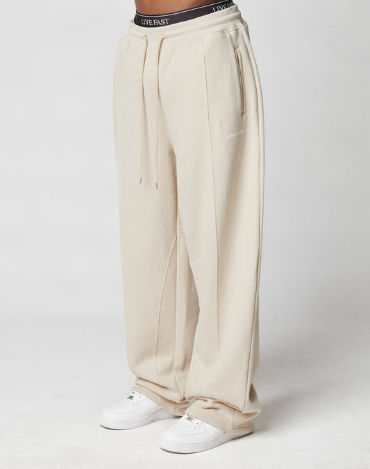 A person wears LFDY Pleated Jogger sweatpants by LIVE FAST DIE YOUNG—loose-fitting, high-waisted, beige with wide fit, elastic waistband, drawstring, side pockets, and “LIVE EAST” on the waist—paired with white sneakers on a white background.| model