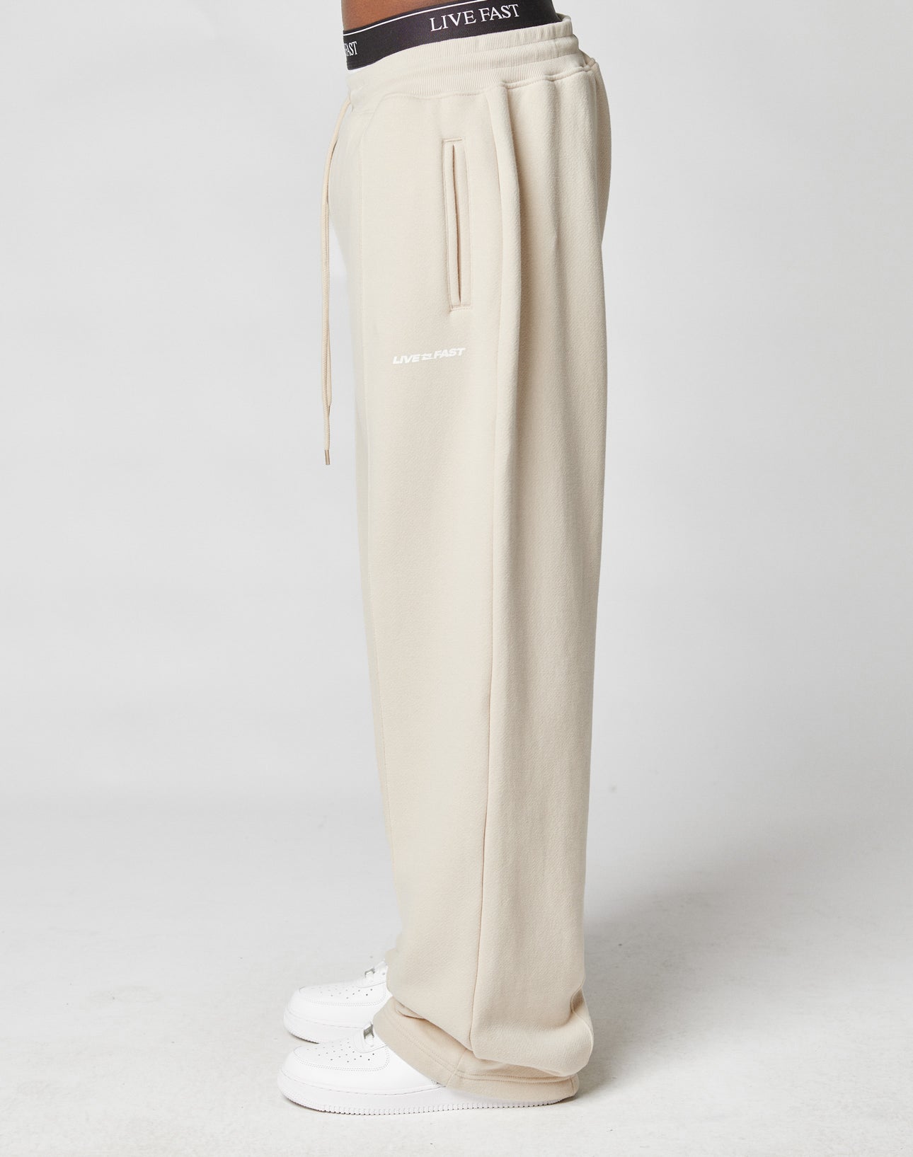A person in beige LFDY Pleated Jogger sweatpants by LIVE FAST DIE YOUNG with LIVE FAST text, a comfy elastic waistband, white sneakers, and black underwear with a LIVE FAST waistband stands sideways against a plain white background.