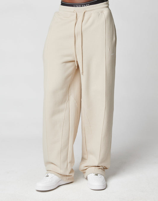A person wearing cream-colored LFDY Pleated Jogger sweatpants by LIVE FAST DIE YOUNG and white sneakers stands against a plain white background, with only the lower half of their body visible.
