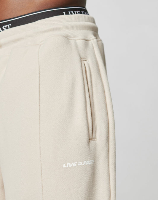 Close-up of a person wearing beige LFDY Pleated Jogger by LIVE FAST DIE YOUNG, featuring a vertical pocket and white LIVE FAST logo. The black elastic waistband has white LIVE FAST text. Only the lower torso and upper legs are shown.
