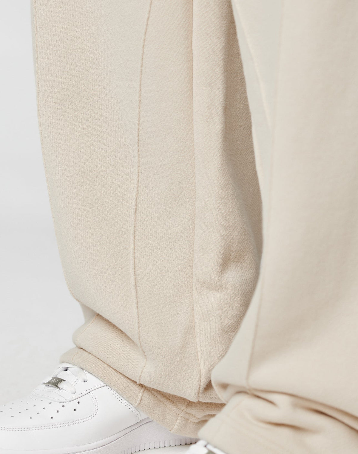 Close-up of lower legs in LIVE FAST DIE YOUNG LFDY Pleated Jogger. The beige, wide fit pants with elastic waistband drape over white sneakers, with soft folds accentuating the textured fabric.