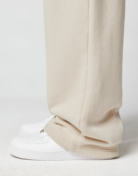 A close-up shows a person wearing LIVE FAST DIE YOUNGs LFDY Pleated Jogger in wide fit beige, featuring an Elastik-Bund, draped over clean white sneakers against a simple white background.