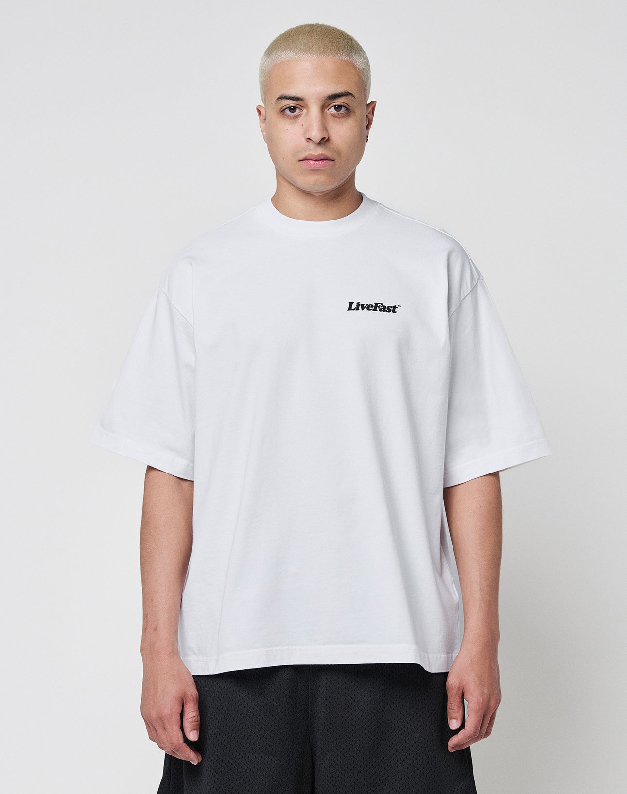 A person with short bleached blond hair wears the LIVE FAST DIE YOUNG Basic 280 Tee, a loose-fitting white 100% Baumwolle shirt with LiveFast in small black letters on the chest, against a plain white background. The oversize fit gives a relaxed vibe.