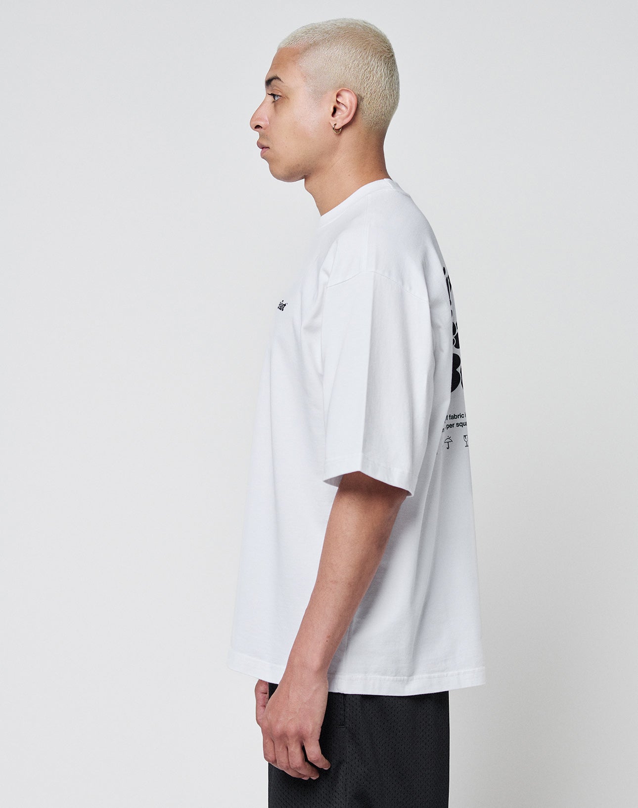 A person with short bleached blonde hair stands in profile, wearing the LIVE FAST DIE YOUNG Basic 280 Tee—an oversize white T-shirt with black front and back print—and black shorts, against a plain light background.