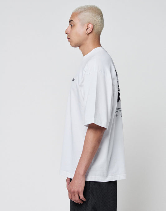 A person with short bleached blonde hair stands in profile, wearing the LIVE FAST DIE YOUNG Basic 280 Tee—an oversize white T-shirt with black front and back print—and black shorts, against a plain light background.