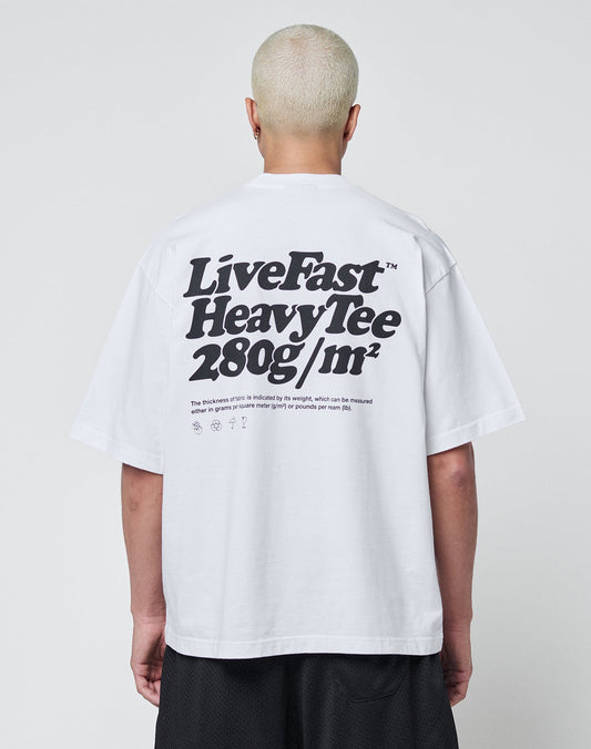 A person with short, bleached hair is seen from behind wearing a white LIVE FAST DIE YOUNG Basic 280 Tee with bold black text and symbols, paired with black shorts.