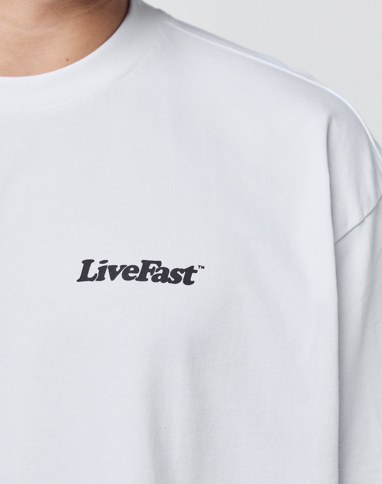 A person wearing the LIVE FAST DIE YOUNG Basic 280 Tee—an oversized, plain white 100% Baumwolle T-shirt with LiveFast in black on the left chest. Only their upper torso and part of their neck are visible.